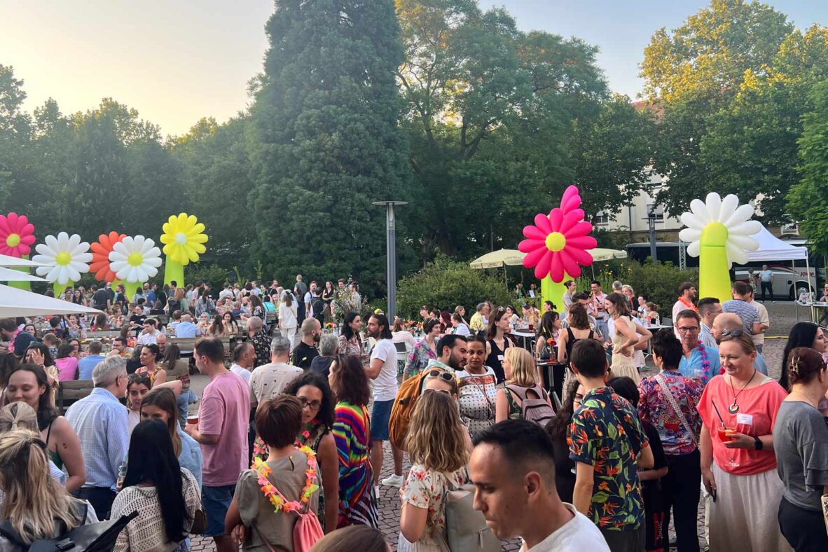 Großes Sommerfest mit vielen Teilnehmenden im Außenbereich. Bunte, aufblasbare Blumen dienen als dekorative Elemente. Die Stimmung ist gesellig und sommerlich.