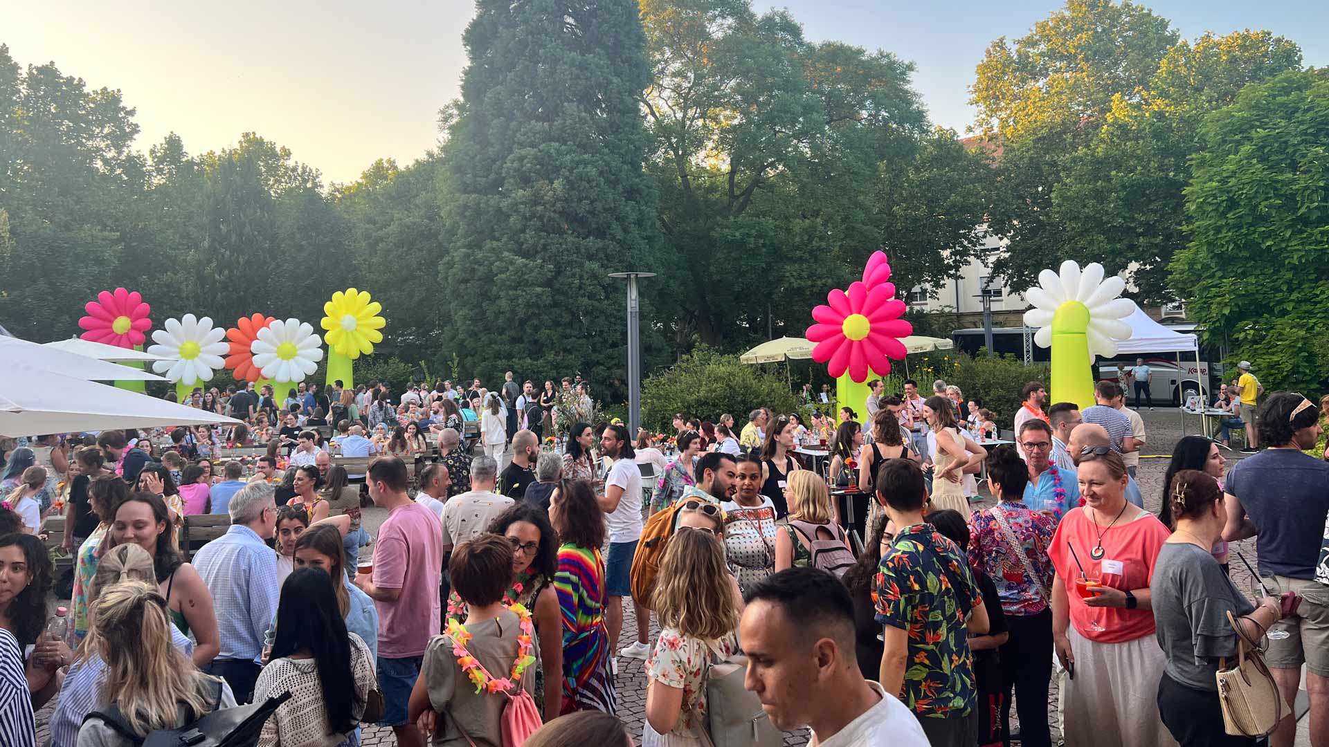 Großes Sommerfest mit vielen Teilnehmenden im Außenbereich. Bunte, aufblasbare Blumen dienen als dekorative Elemente. Die Stimmung ist gesellig und sommerlich.